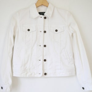 Pode incluir: Casaco de ganga branco com gola clássica, fecho de botões e dois bolsos no peito. O casaco tem mangas compridas e botões castanhos escuros. Está pendurado num cabide de madeira.