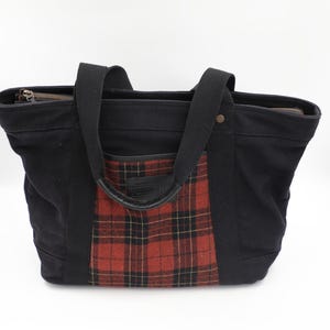 Puede incluir: Un bolso tote negro con un panel central de cuadros rojos y negros en la parte frontal. El bolso cuenta con dos asas de tela negra, un cierre de cremallera superior y un peque&ntilde;o detalle de remache met&aacute;lico en la esquina superior derecha.
