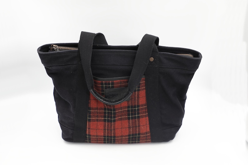 Puede incluir: Un bolso tote negro con un panel central de cuadros rojos y negros en la parte frontal. El bolso cuenta con dos asas de tela negra, un cierre de cremallera superior y un peque&ntilde;o detalle de remache met&aacute;lico en la esquina superior derecha.