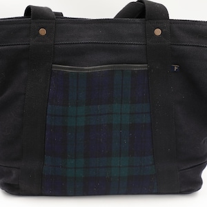 Puede incluir: Un bolso tote de lona negra con un bolsillo central de estampado de cuadros en azul oscuro y verde. El bolso tiene dos asas de tela negra resistentes sujetas con remaches de color cobre y una peque&ntilde;a etiqueta con la letra P en el borde.
