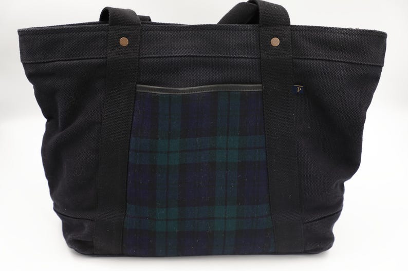 Puede incluir: Un bolso tote de lona negra con un bolsillo central de estampado de cuadros en azul oscuro y verde. El bolso tiene dos asas de tela negra resistentes sujetas con remaches de color cobre y una peque&ntilde;a etiqueta con la letra P en el borde.