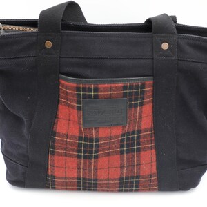 Puede incluir: Un bolso tote de lona negra con un bolsillo frontal de cuadros rojos y negros y un parche rectangular de cuero negro. El bolso incluye dos asas de tela negra, remaches de color cobre y un cierre de cremallera en la parte superior.