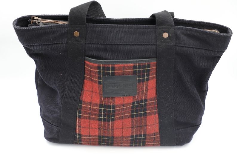 Puede incluir: Un bolso tote de lona negra con un bolsillo frontal de cuadros rojos y negros y un parche rectangular de cuero negro. El bolso incluye dos asas de tela negra, remaches de color cobre y un cierre de cremallera en la parte superior.