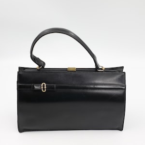 Puede incluir: Un bolso de mano negro estructurado con un asa superior única y un cierre de metal en tono dorado. La parte frontal presenta un detalle de correa horizontal con una pequeña hebilla ovalada en tono dorado. El bolso tiene forma rectangular.