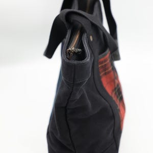 Puede incluir: Un bolso tote de lona negra con un panel lateral de patr&oacute;n de cuadros escoceses en rojo y negro. El bolso incluye asas de tela negra, un cierre de cremallera superior con tirador met&aacute;lico y una base estructurada, capturado desde un &aacute;ngulo lateral sobre un fondo blanco.