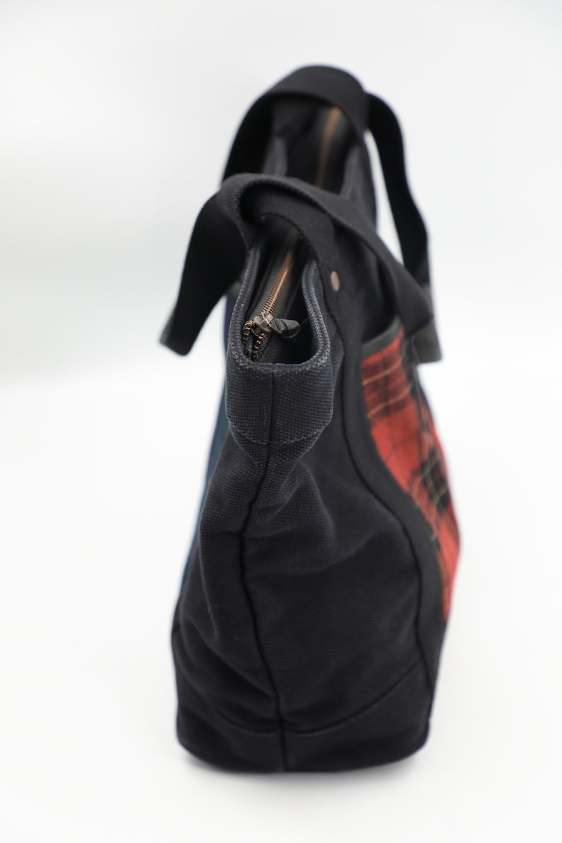 Puede incluir: Un bolso tote de lona negra con un panel lateral de patr&oacute;n de cuadros escoceses en rojo y negro. El bolso incluye asas de tela negra, un cierre de cremallera superior con tirador met&aacute;lico y una base estructurada, capturado desde un &aacute;ngulo lateral sobre un fondo blanco.