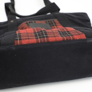 Puede incluir: Un bolso tote de lona negra que presenta un bolsillo frontal rectangular con un patr&oacute;n de cuadros en rojo, negro y amarillo. El bolso incluye dos asas de tela negra unidas a la parte superior, una base reforzada y una estructura de tela oscura.