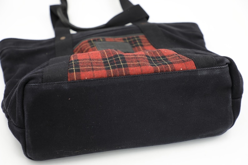 Puede incluir: Un bolso tote de lona negra que presenta un bolsillo frontal rectangular con un patr&oacute;n de cuadros en rojo, negro y amarillo. El bolso incluye dos asas de tela negra unidas a la parte superior, una base reforzada y una estructura de tela oscura.