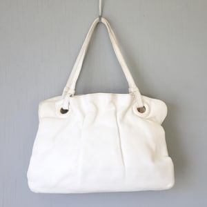 Puede incluir: Un bolso tote de cuero blanco con dos asas y detalles metálicos dorados. El bolso cuelga sobre un fondo gris. El bolso tote tiene un diseño suave y holgado.