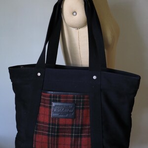 Puede incluir: Un bolso tote de lona negra con un panel central de tela a cuadros rojos, negros y amarillos. El bolso tiene dos correas negras para el hombro, remaches plateados y un parche de cuero negro con el nombre de la marca Pendleton en relieve.