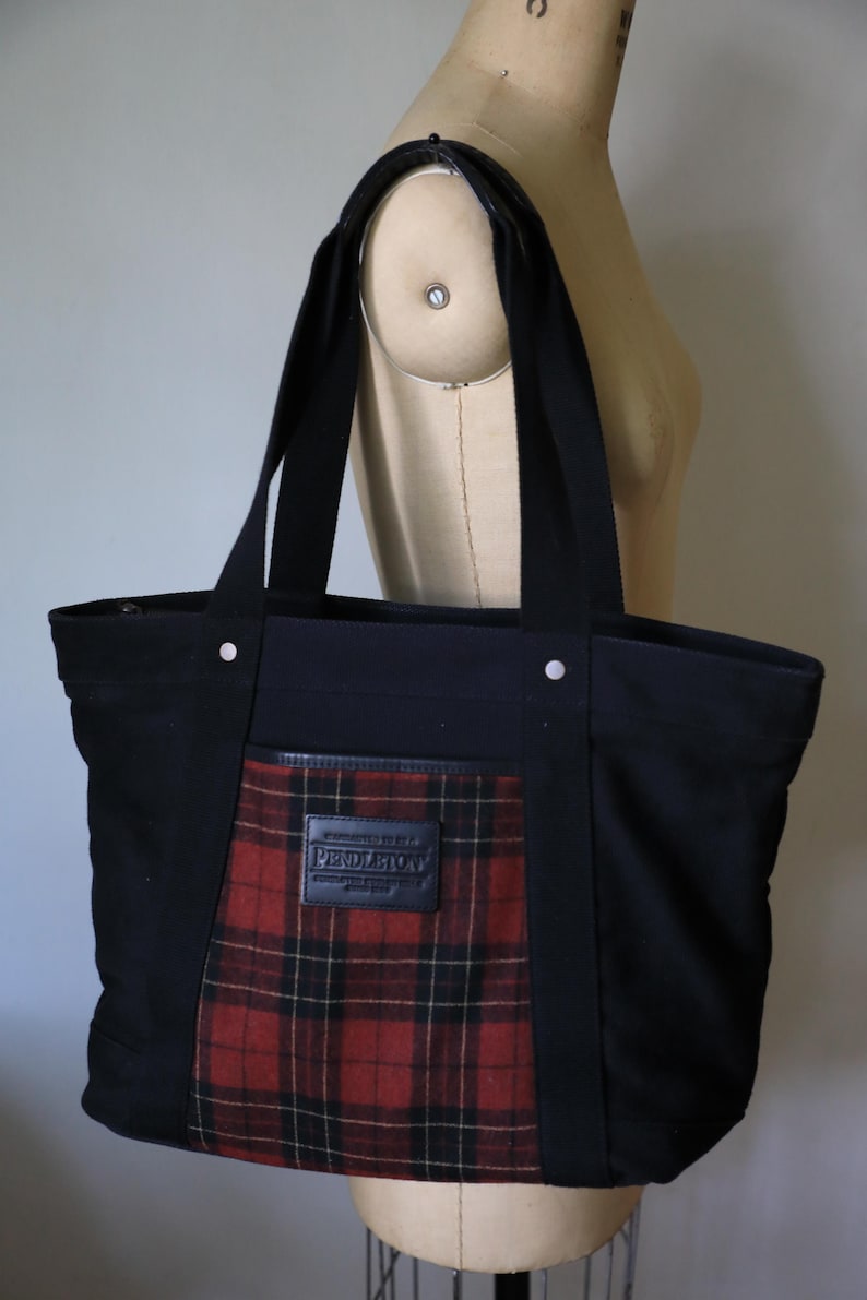 Puede incluir: Un bolso tote de lona negra con un panel central de tela a cuadros rojos, negros y amarillos. El bolso tiene dos correas negras para el hombro, remaches plateados y un parche de cuero negro con el nombre de la marca Pendleton en relieve.