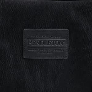 May include: A black canvas tote bag featuring a rectangular black leather patch embossed with the text Pendleton, Warranted To Be A Pendleton, Pendleton Woolen Mills, Since 1863. The bag includes black fabric straps secured with silver metal rivets.