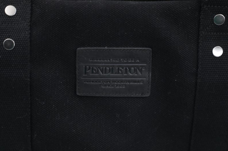 May include: A black canvas tote bag featuring a rectangular black leather patch embossed with the text Pendleton, Warranted To Be A Pendleton, Pendleton Woolen Mills, Since 1863. The bag includes black fabric straps secured with silver metal rivets.