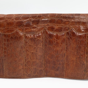 May include: A brown leather clutch bag with a crocodile skin effect. The rectangular bag features a scalloped flap and a curved base. The leather is a deep, warm brown, and the texture is detailed and prominent.