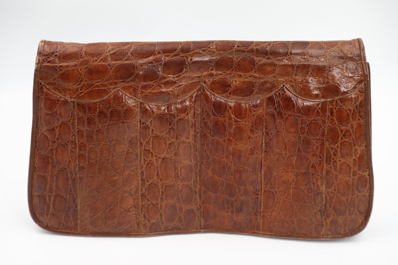 May include: A brown leather clutch bag with a crocodile skin effect. The rectangular bag features a scalloped flap and a curved base. The leather is a deep, warm brown, and the texture is detailed and prominent.