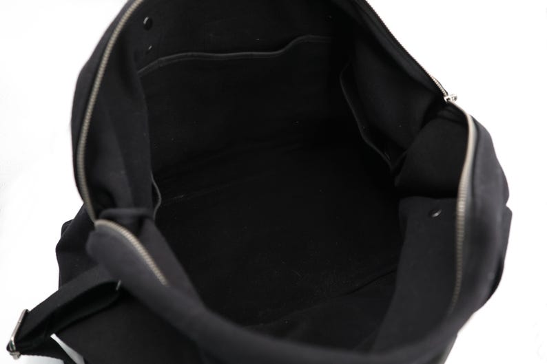 May include: A black bag with a wide open top, featuring a silver-toned metal zipper closure that runs along the upper edge. The interior is lined with black fabric and includes a flat pocket on the back wall and a snap button fastener.