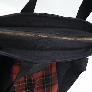 Puede incluir: Un bolso tote de lona negra con cierre de cremallera superior y riel met&aacute;lico. El exterior del bolso incluye un parche rectangular con un patr&oacute;n de cuadros en rojo, negro y amarillo. El bolso tiene dos asas de tela negra incorporadas.
