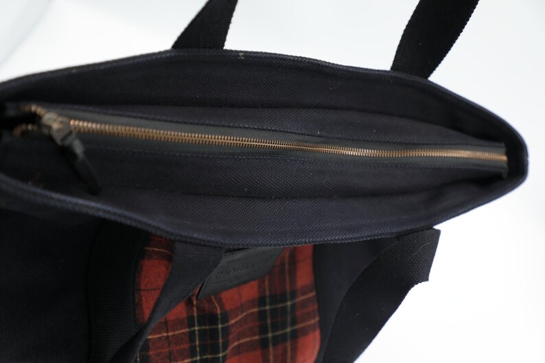 Puede incluir: Un bolso tote de lona negra con cierre de cremallera superior y riel met&aacute;lico. El exterior del bolso incluye un parche rectangular con un patr&oacute;n de cuadros en rojo, negro y amarillo. El bolso tiene dos asas de tela negra incorporadas.