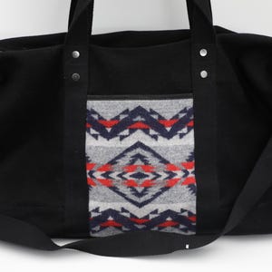 May include: A black duffel bag featuring a central panel with a geometric pattern in shades of gray, navy blue, and red. The bag includes black fabric handles, silver-toned metal rivets, a top zipper closure, and a detachable black shoulder strap.