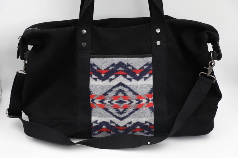 May include: A black duffel bag featuring a central panel with a geometric pattern in shades of gray, navy blue, and red. The bag includes black fabric handles, silver-toned metal rivets, a top zipper closure, and a detachable black shoulder strap.