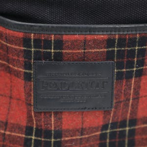 Puede incluir: Un bolso de lona negro con un bolsillo frontal con un patr&oacute;n de lana a cuadros rojos y negros. Un parche rectangular de cuero negro est&aacute; centrado en el bolsillo, grabado con el texto Pendleton, Warranted to be a Pendleton, y Since 1863.