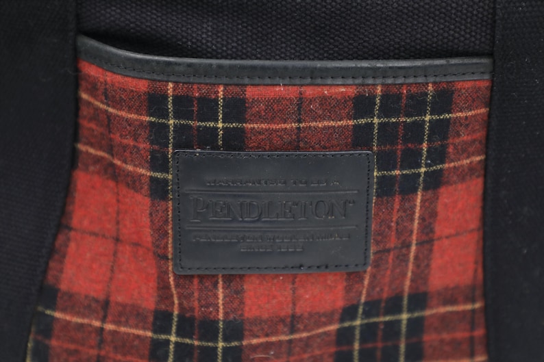 Puede incluir: Un bolso de lona negro con un bolsillo frontal con un patr&oacute;n de lana a cuadros rojos y negros. Un parche rectangular de cuero negro est&aacute; centrado en el bolsillo, grabado con el texto Pendleton, Warranted to be a Pendleton, y Since 1863.