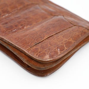 May include: Close-up of a brown leather clutch bag with a crocodile skin texture. The bag is rectangular with rounded corners and a flap closure. The leather has a rich, warm tone.