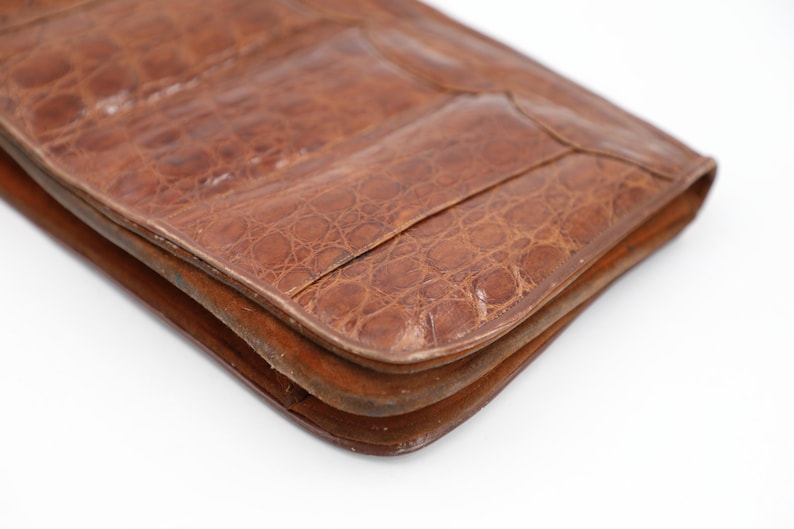 May include: Close-up of a brown leather clutch bag with a crocodile skin texture. The bag is rectangular with rounded corners and a flap closure. The leather has a rich, warm tone.