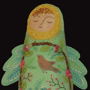 May include: A handmade felt doll with a serene face, wearing a yellow headdress and adorned with gold beads. The doll's body is light green, decorated with a brown bird, pink flowers, and embroidered leaves. It has light green wings with detailed stitching.
