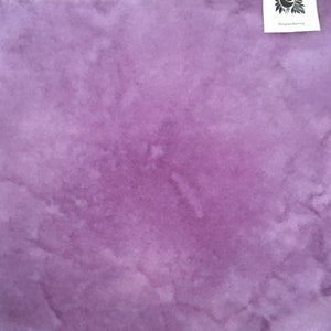 May include: A close-up of a piece of fabric in shades of purple. The fabric has a watercolor-like effect with lighter and darker areas, creating a textured appearance. A small white tag in the upper right corner reads "Boysenberry".