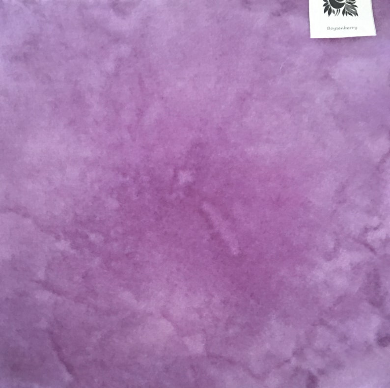 May include: A close-up of a piece of fabric in shades of purple. The fabric has a watercolor-like effect with lighter and darker areas, creating a textured appearance. A small white tag in the upper right corner reads "Boysenberry".