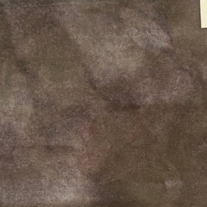 May include: A close-up of a fabric with a mottled appearance, featuring shades of brown, gray, and hints of purple. The texture appears soft and slightly distressed. A small tag in the upper right corner reads "Pine Cones".