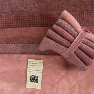 May include: A collection of mauve-colored fabric swatches with various textures, including houndstooth and solid patterns. A stack of folded fabric is secured with a matching ribbon. A tag reads "Olympic Wool Works, Port Angeles, WA".