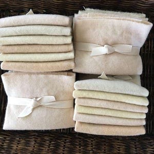 May include: Stacks of off-white and beige fabric squares and rectangles are arranged in a woven basket. Some stacks are tied with a cream-colored ribbon. The fabrics vary in shade from light cream to a deeper beige.