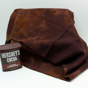 May include: A folded, textured, dark brown fabric with lighter brown patches, next to a Hershey's Cocoa container. The container is dark brown with white text that reads "HERSHEY'S COCOA 100% CACAO NATURAL UNSWEETENED".