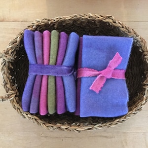 May include: A woven basket holds two bundles of colorful felt squares. One bundle is wrapped with a purple band, showcasing shades of purple, pink, and green. The other bundle is tied with a pink ribbon, displaying a solid purple hue.