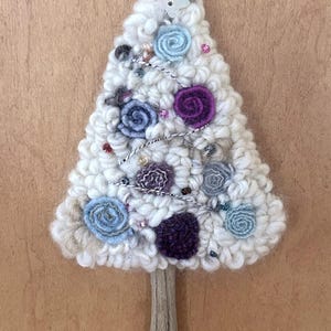 May include: A handcrafted Christmas tree decoration made of white, textured yarn. It is adorned with blue and purple yarn flowers, beads, and a star-shaped button at the top. The tree is mounted on a wooden stick.