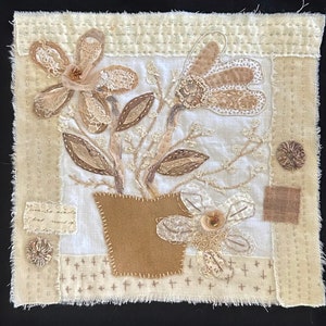 May include: A textile art piece featuring a floral design in shades of beige and brown. The artwork includes appliqued flowers, leaves, and a pot, with decorative stitching and a frayed edge. The piece has a vintage, handmade aesthetic.