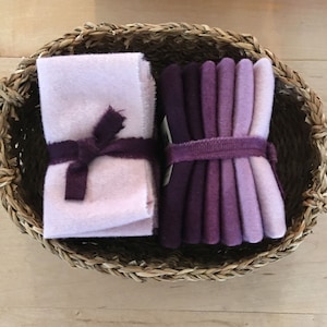 May include: An oval woven basket holds two bundles of felt fabric. One bundle is light pink and tied with a dark purple ribbon. The other bundle contains shades of purple felt, secured with a matching purple band. The basket has two handles.