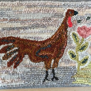May include: Hand-hooked rug featuring a brown turkey with a red wattle and black legs. The turkey stands next to a pink flower and green leaves. The background is a mix of gray, beige, and brown.