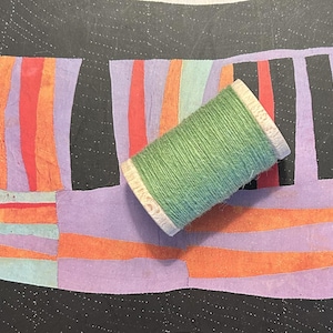 May include: A green thread spool rests on a colorful patchwork textile. The textile features a black background with a patchwork design of purple, orange, and red vertical stripes. The thread spool is light green.