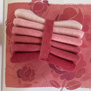 May include: A bundle of felt squares in varying shades of pink and red, secured with a matching fabric band. The felt colors range from pale pink to deep red. The background features a floral pattern in similar tones.