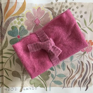 May include: A bright pink, rectangular felt pouch with a matching bow closure. The pouch is made of a soft, textured material. The background features a watercolor painting with floral and botanical elements.