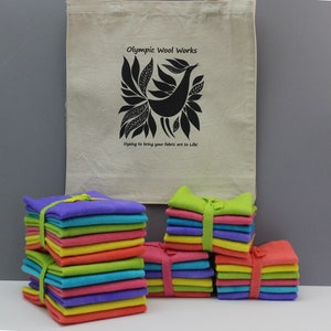 May include: Stacks of colorful fabric squares tied with ribbons in shades of purple, blue, green, pink, orange, and yellow. A beige tote bag with a black bird design and the text "Olympic Wool Works" is in the background.