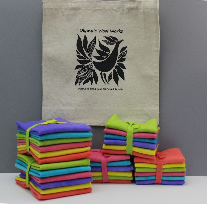 May include: Stacks of colorful fabric squares tied with ribbons in shades of purple, blue, green, pink, orange, and yellow. A beige tote bag with a black bird design and the text "Olympic Wool Works" is in the background.