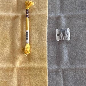 May include: A close-up shot of embroidery supplies. A yellow embroidery floss is on the left, and a gray thread spool is on the right. The background is divided into two sections, one yellow and one gray.
