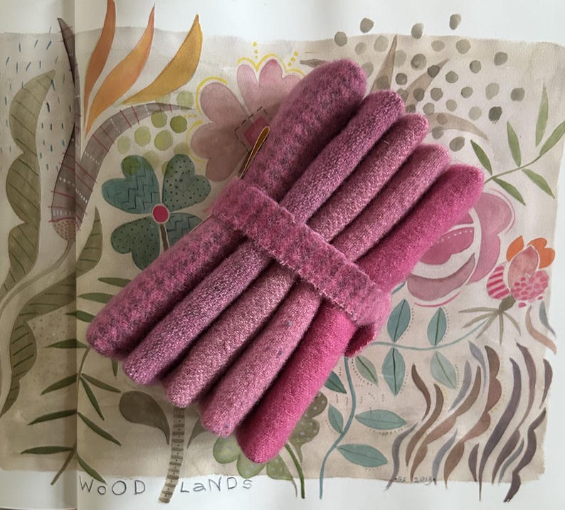 May include: A bundle of pink felted wool tubes, bound together with a matching strap. The tubes vary in shade from light to deep pink. The background features a watercolor illustration with floral and botanical elements.