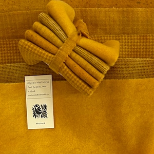 May include: Mustard-colored fabric bundle with various textures, including solid, dotted, and houndstooth patterns. A label from Olympic Wool Works is visible, with the word "Mustard" printed on it. The fabrics are tied together with a matching band.