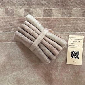 May include: A bundle of ecru-colored fabric swatches, bound with a matching band. The swatches display varying shades of beige and off-white. A small tag with the text "Ecru" and a logo is visible.