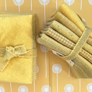 May include: Two stacks of yellow fabric squares, one tied with a bow and the other with a woven band. The fabric has a soft, textured appearance. Some squares have a houndstooth pattern. The background is a yellow pattern with white circles.